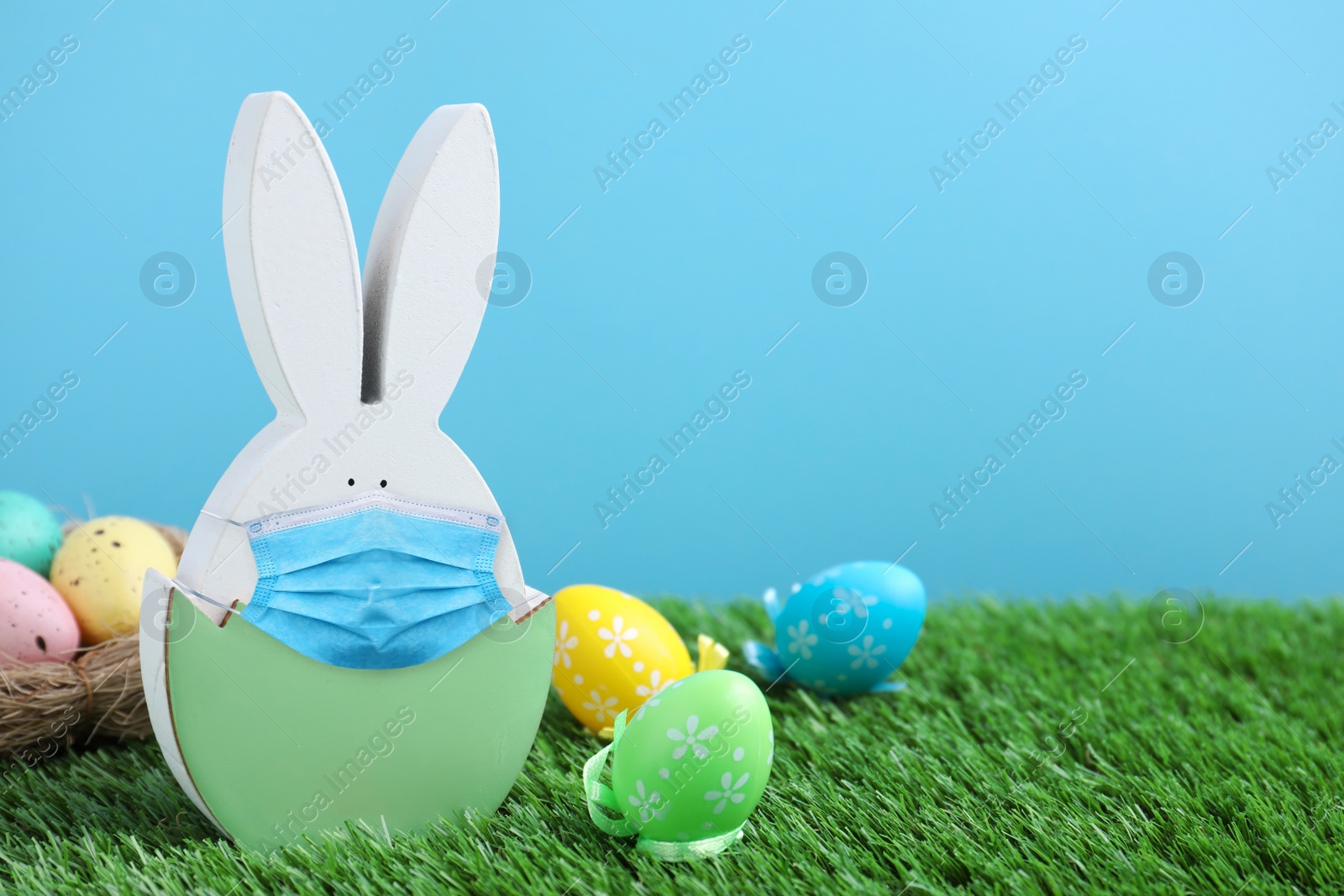Image of COVID-19 pandemic. Easter bunny figure in protective mask and dyed eggs on green grass against light blue background, space for text