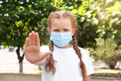 Photo of Little girl in protective mask showing stop gesture outdoors. Prevent spreading of coronavirus