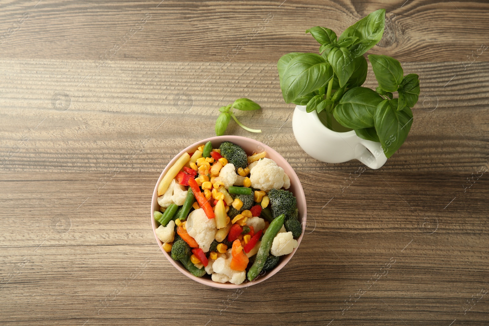 Photo of Mix of different frozen vegetables in bowl and basil on wooden table, flat lay