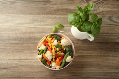 Mix of different frozen vegetables in bowl and basil on wooden table, flat lay