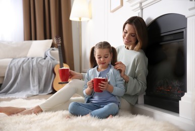 Photo of Happy woman and her daughter with cups of hot drink resting near fireplace at home