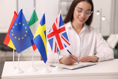 Photo of Young woman writing in notebook at white table indoors, focus on different flags