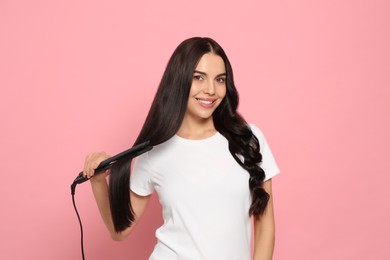 Photo of Beautiful happy woman using hair iron on pink background