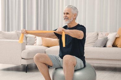 Senior man doing exercise with elastic resistance band on fitness ball at home