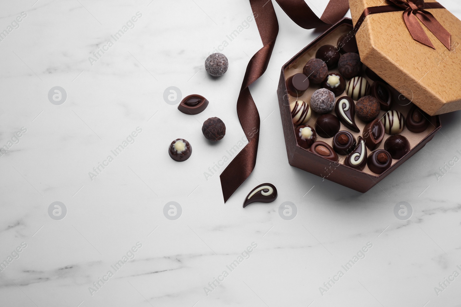 Photo of Open box of delicious chocolate candies and brown ribbon on white marble table, flat lay. Space for text