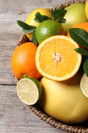 Photo of Different fresh citrus fruits and leaves in wicker basket on wooden table, closeup