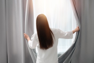 Photo of Woman opening window curtains at home, back view. Lazy morning