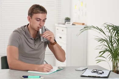 Young man with glass of water writing in notebook at table indoors