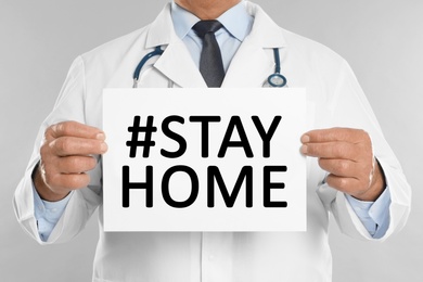 Senior doctor holding paper with hashtag Stayhome on white background, closeup. Protective measure during coronavirus pandemic