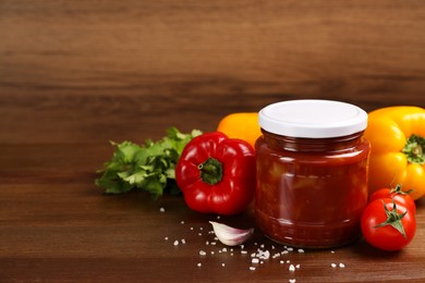 Glass jar of delicious canned lecho and fresh ingredients on wooden table. Space for text