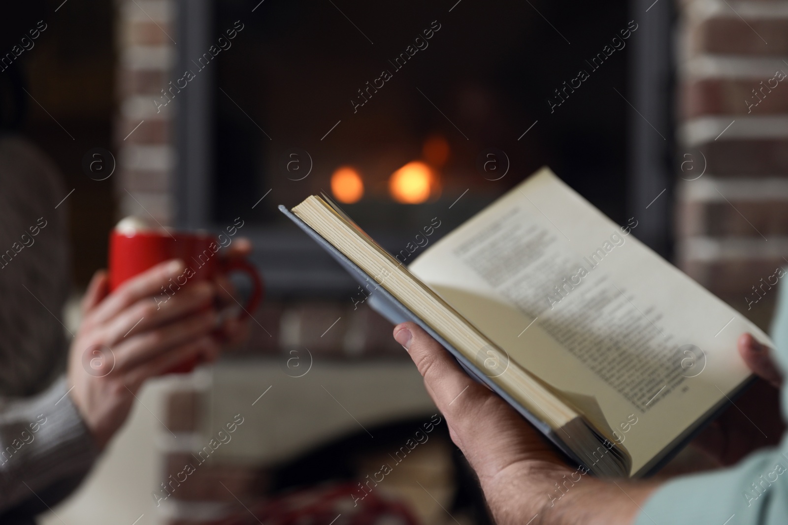 Photo of Man reading book and his girlfriend near burning fireplace at home, closeup