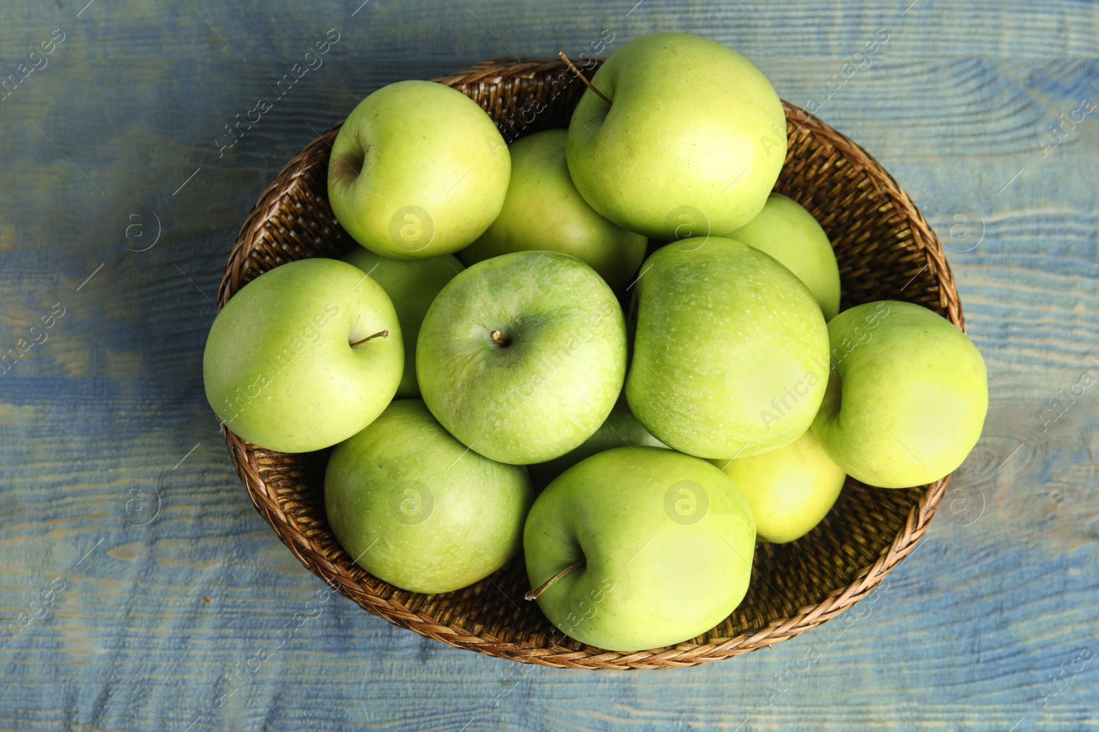 Photo of Wicker bowl of fresh ripe green apples on blue wooden table, top view