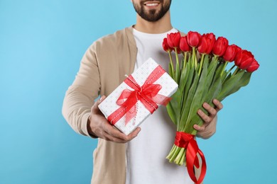 Photo of Happy man with red tulip bouquet and gift box on light blue background, closeup. 8th of March celebration