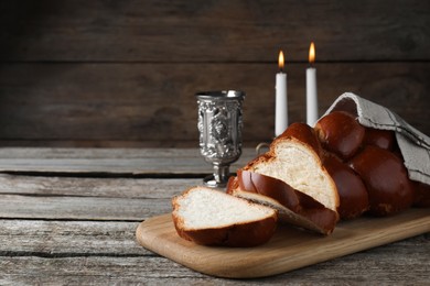 Photo of Cut homemade braided bread, goblet and candles on wooden table, space for text. Traditional Shabbat challah