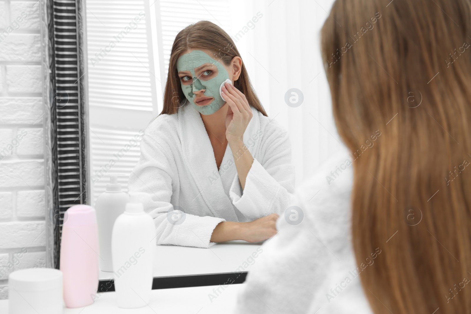 Photo of Beautiful woman removing homemade clay mask from her face at mirror indoors