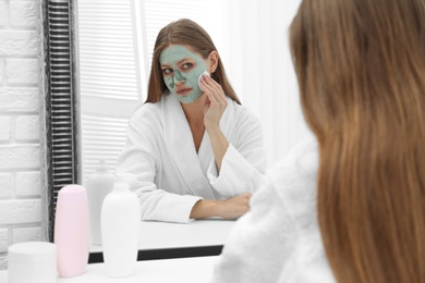 Beautiful woman removing homemade clay mask from her face at mirror indoors