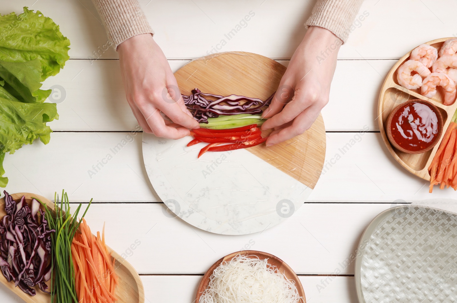 Photo of Making delicious spring rolls. Woman wrapping fresh vegetables into rice paper at white wooden table, top view