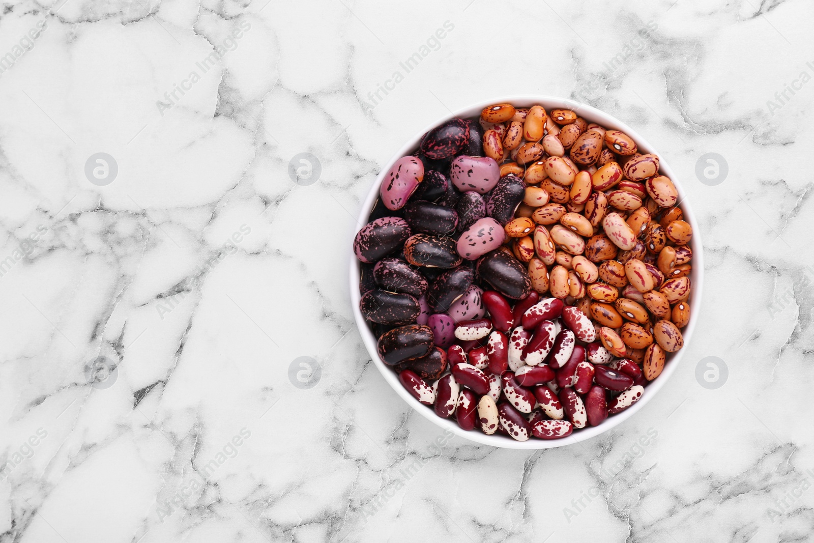 Photo of Different kinds of dry kidney beans in bowl on white marble table, top view