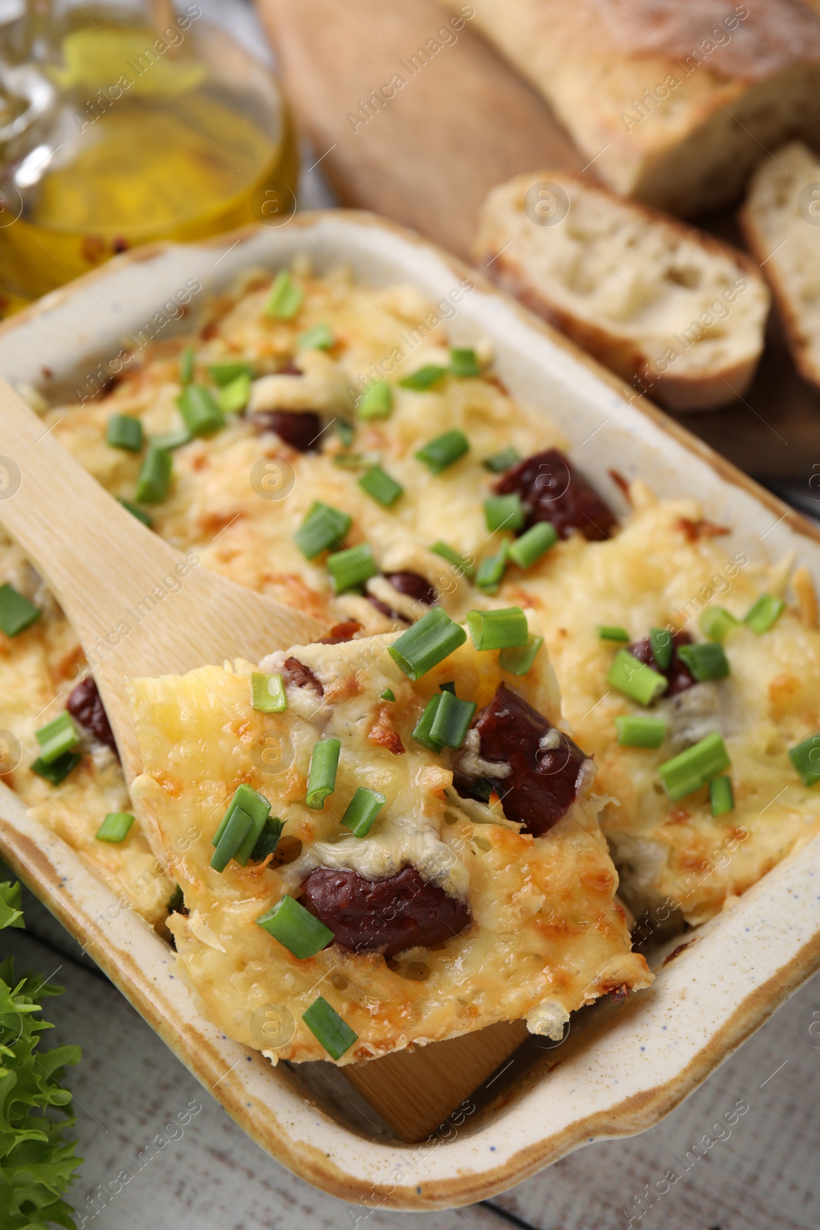 Photo of Taking piece of tasty sausage casserole from baking dish at white table, closeup