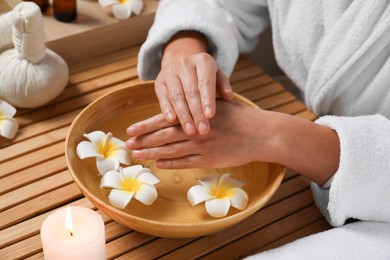 Woman soaking her hands in bowl of water and flowers at wooden table, closeup. Spa treatment