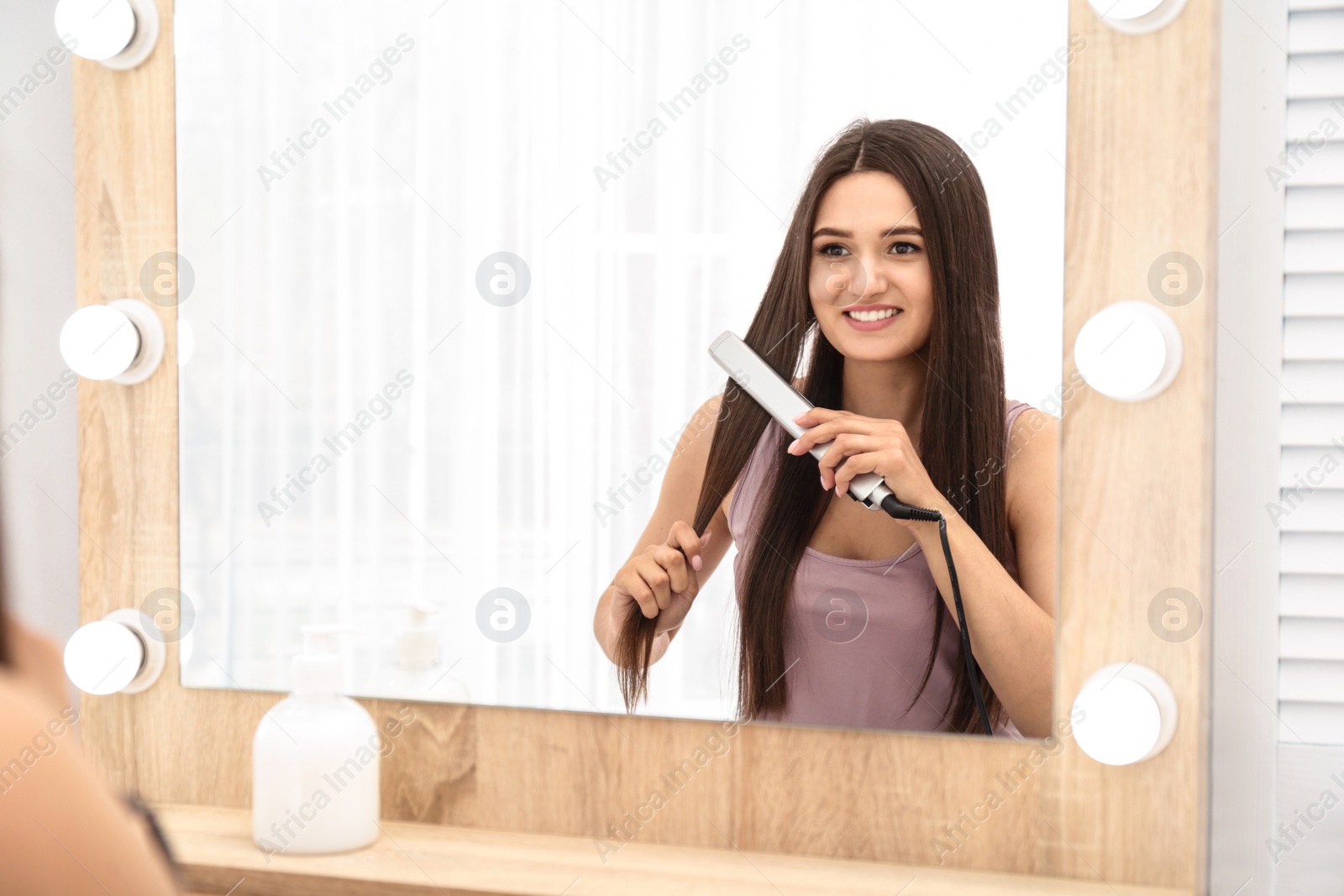 Photo of Young woman using hair iron near mirror indoors, space for text