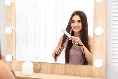 Young woman using hair iron near mirror indoors, space for text