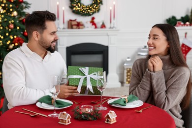 Photo of Happy young man presenting Christmas gift to his girlfriend at table indoors