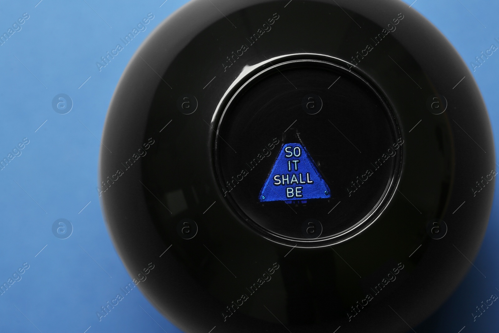Photo of Magic eight ball with prediction So It Shall Be on blue background, above view