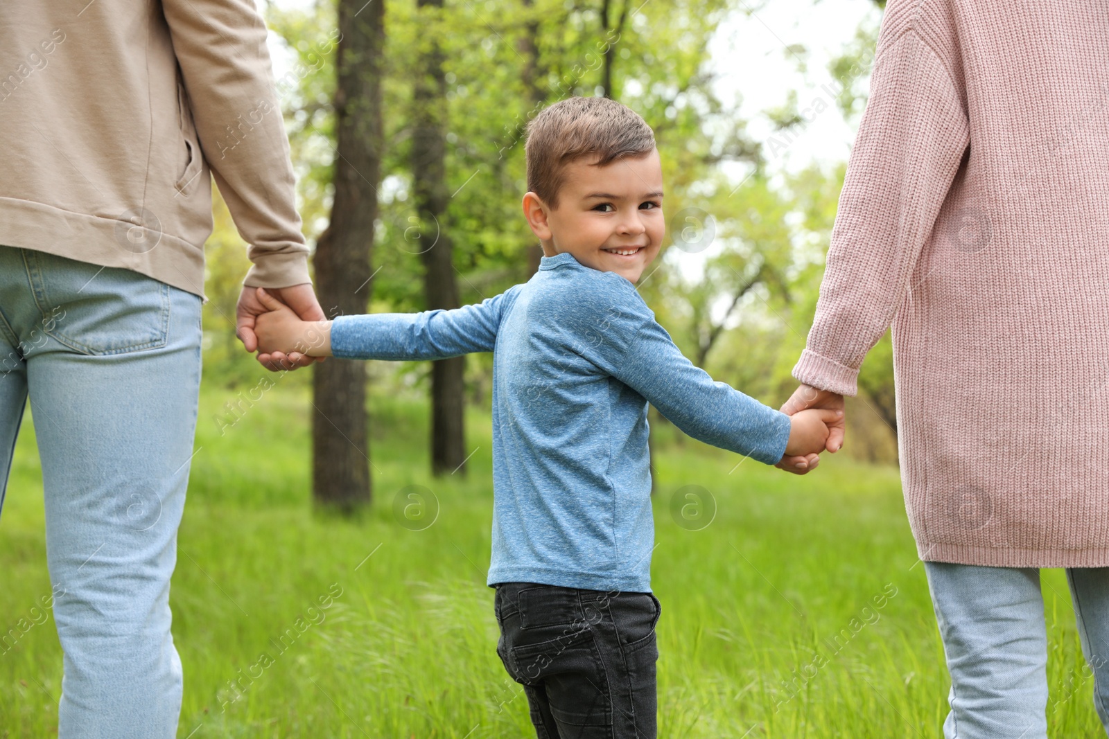 Photo of Happy little child holding hands with his parents in park. Family weekend