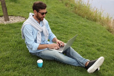 Man in sunglasses with laptop and coffee on green grass near lake