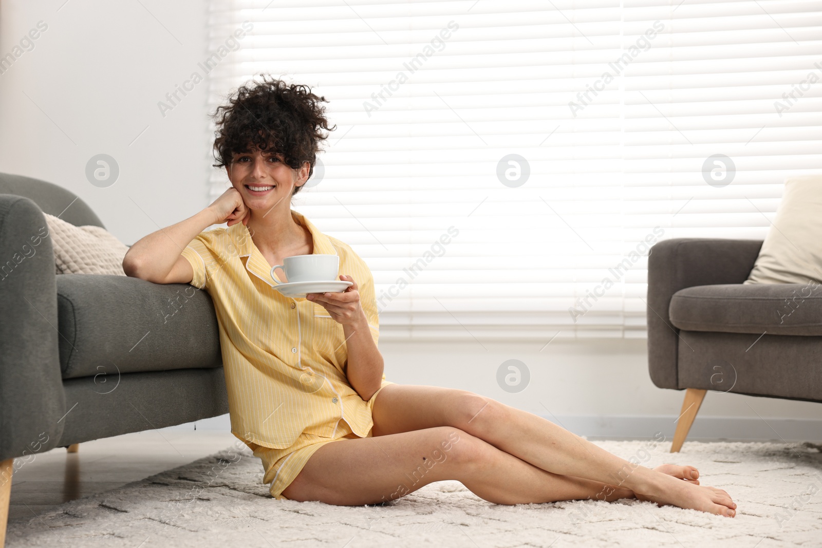 Photo of Beautiful young woman in stylish pyjama with cup of drink on floor at home