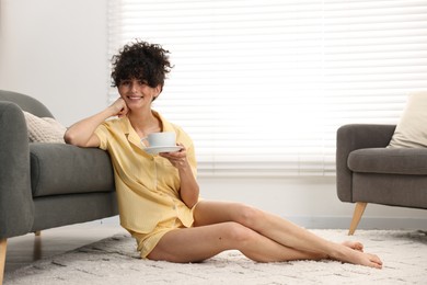 Photo of Beautiful young woman in stylish pyjama with cup of drink on floor at home