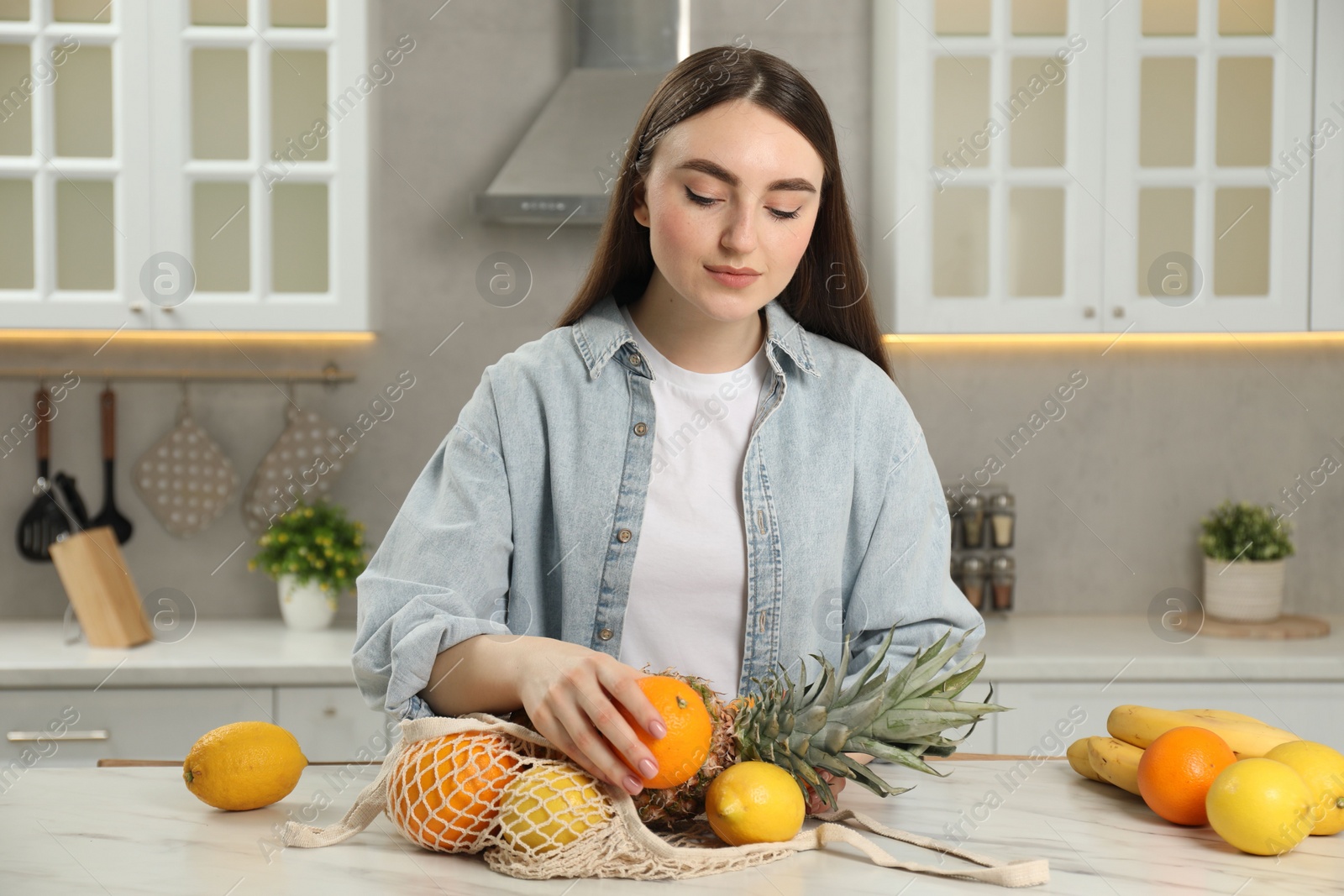 Photo of Woman with string bag of fresh fruits at light marble table in kitchen