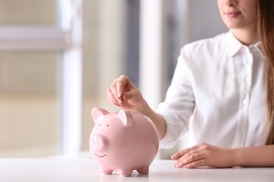 Photo of Woman putting coin into piggy bank at table indoors, closeup. Space for text