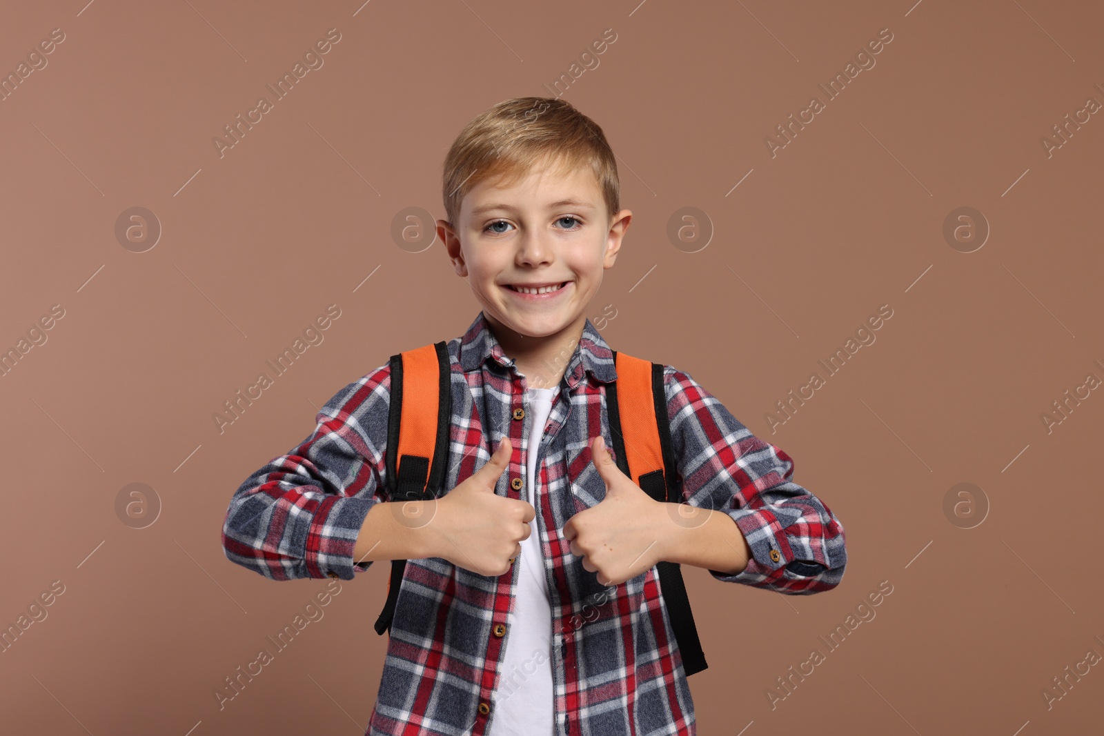 Photo of Happy schoolboy with backpack showing thumbs up gesture on brown background