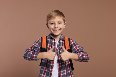 Happy schoolboy with backpack showing thumbs up gesture on brown background