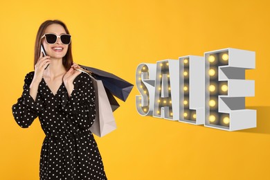 Happy young woman with shopping bags and smartphone on yellow background. Lamp in shape of word Sale