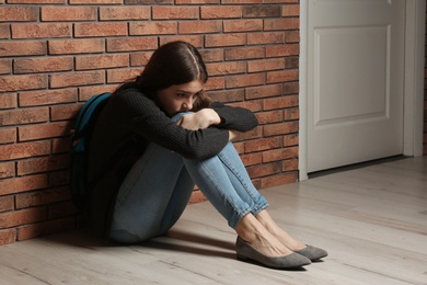Upset teenage girl with backpack sitting on floor near brick wall