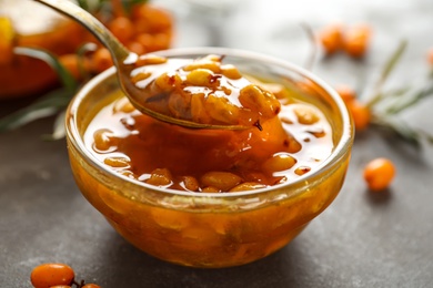 Photo of Spoon with delicious sea buckthorn jam over bowl on grey table, closeup