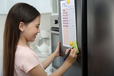 Photo of Little girl drawing funny face on note near to do list in kitchen