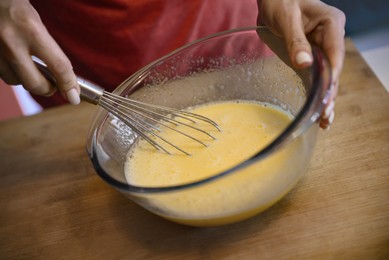 Photo of Woman whisking eggs for crepe batter in glass bowl on wooden table, closeup