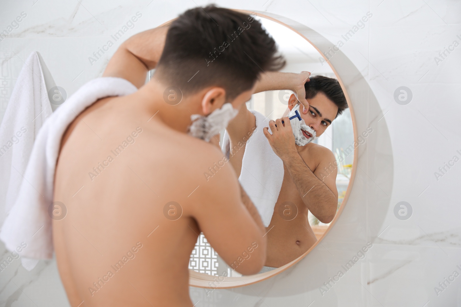 Photo of Handsome young man shaving with razor near mirror in bathroom