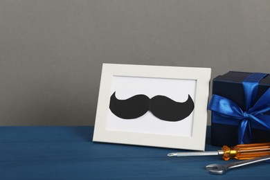 Photo of Happy Father's Day. Gift box, tools and frame with mustache on blue wooden table, closeup. Space for text