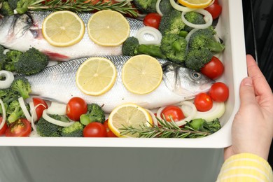 Photo of Woman putting baking dish with raw fish and vegetables into oven, top view