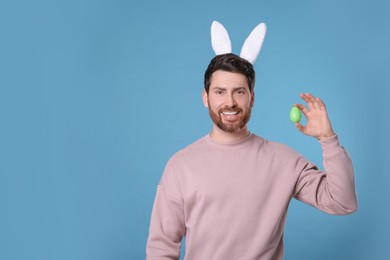 Happy man in cute bunny ears headband covering eye with Easter egg on light blue background. Space for text