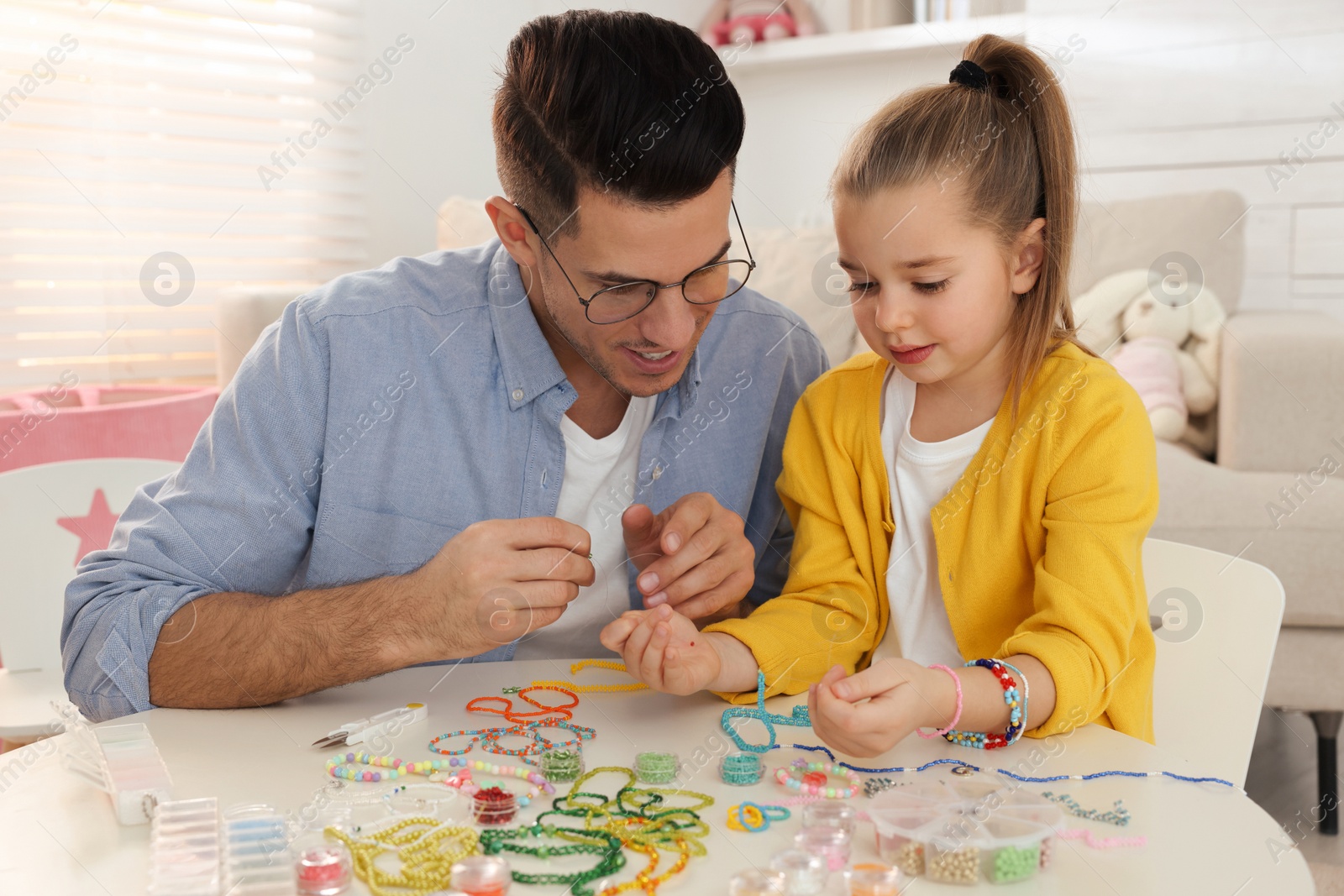 Photo of Happy father with his cute daughter making beaded jewelry at table in room