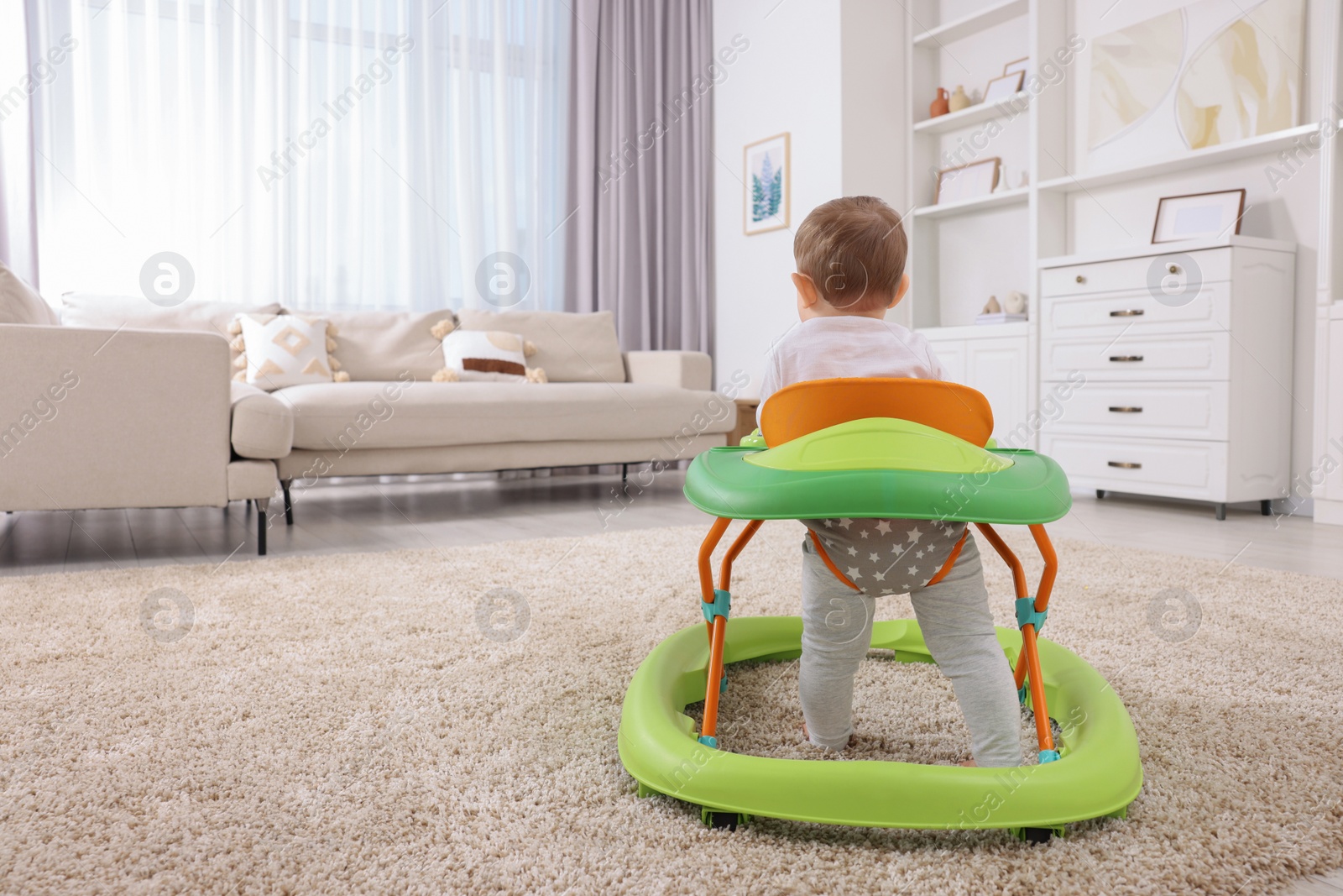 Photo of Cute baby making first steps with toy walker at home, back view. Space for text