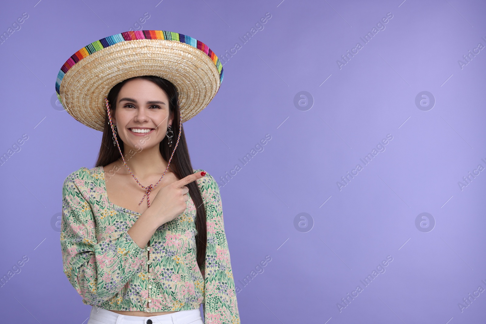 Photo of Young woman in Mexican sombrero hat pointing at something on violet background. Space for text