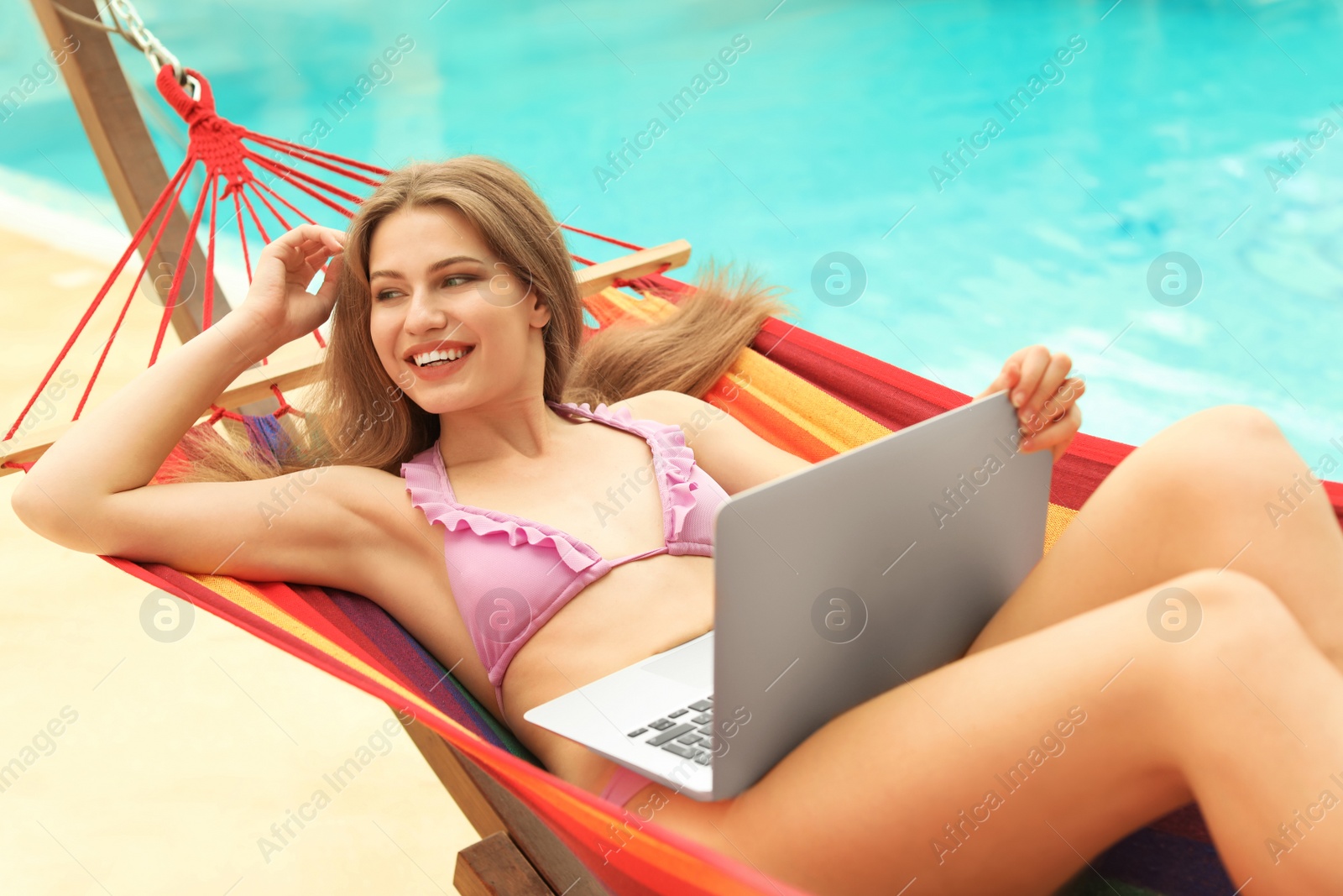 Photo of Young woman with laptop resting in hammock near pool