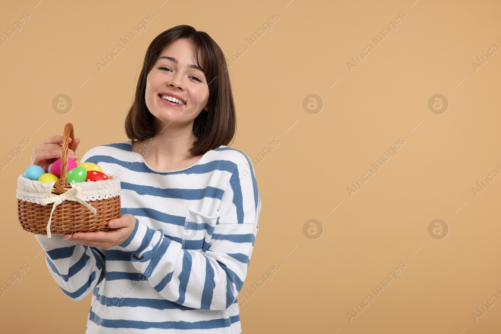 Photo of Easter celebration. Happy woman with wicker basket full of painted eggs on beige background, space for text